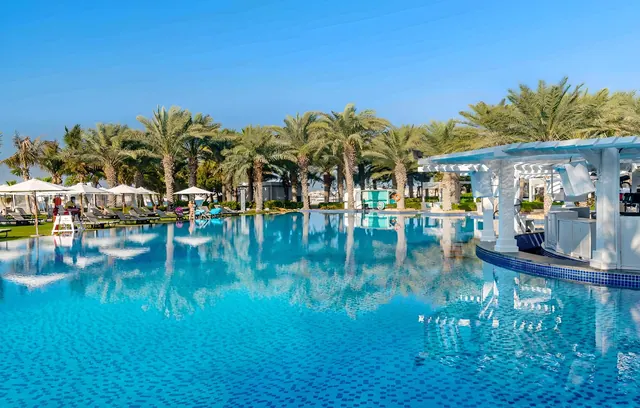 A large blue swimming pool with a white pool bar on the right, white parasols and sun loungers beside palm trees under a clear sky.