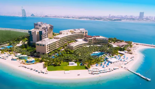 Aerial view of a beachfront resort with a sandy shoreline, a pool, rows of sun loungers and umbrellas, and the Burj Al Arab visible in the distance across the water.