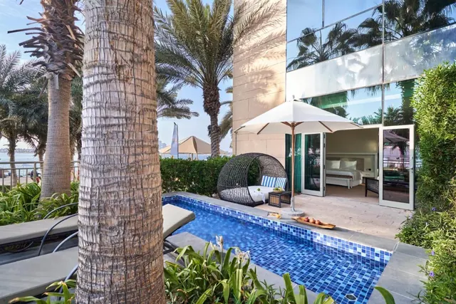 A small private plunge pool sits on a terrace beside a wicker daybed, a white umbrella and sun loungers, with an open glass door leading to a bedroom.