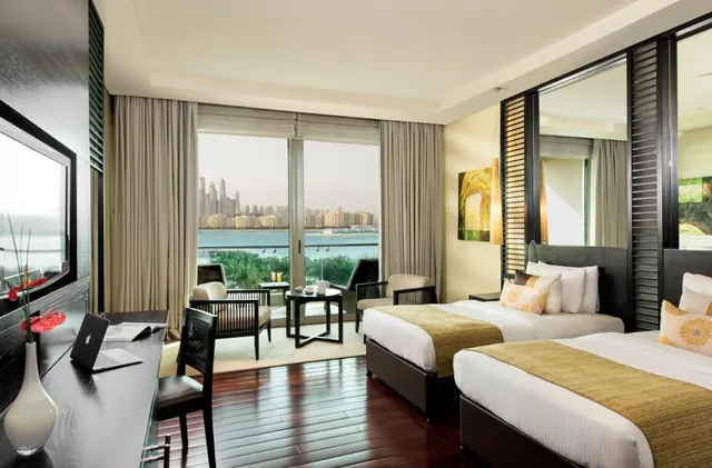A twin-bedded hotel room with a desk, armchairs and floor-to-ceiling glass doors opening onto a balcony with a view of the sea and a city skyline.