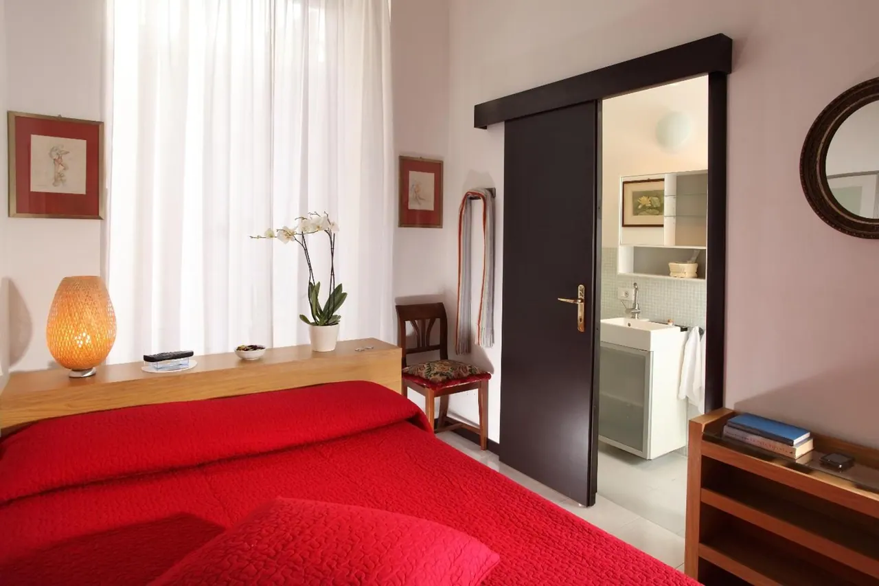 A bedroom with a red bedspread, a wooden headboard shelf holding a lamp and an orchid, and an open sliding door showing a bathroom sink.