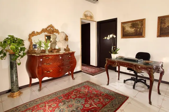 A room with an ornate wooden commode and gilt mirror, a writing desk with a laptop and office chair, and a red patterned rug on a tiled floor.