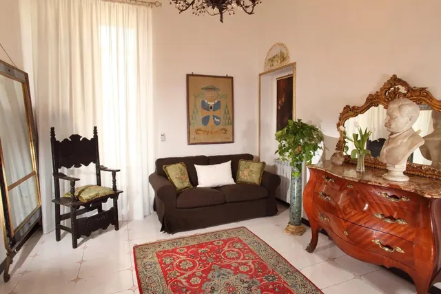 A living room with a dark brown sofa, a carved wooden armchair, an ornate marble-topped commode with a bust and mirror, a potted plant, and a red patterned rug.
