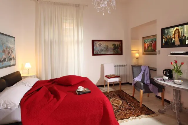 A bedroom with a double bed under a red quilt, a seating chair, a wall-mounted television and a round table with a kettle and tulips.