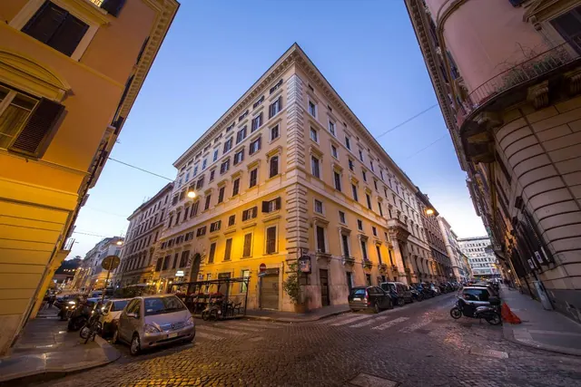 A wide-angle dusk view of a lit yellow corner building on a cobbled Roman street, with parked cars and scooters along the road.