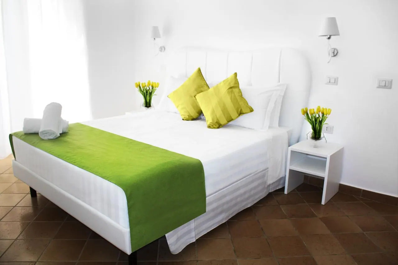 A made double bed with white linens, green and yellow cushions and a green runner, with yellow tulips on two white side tables against white walls and a terracotta tile floor.