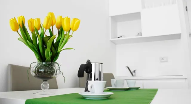 A white kitchenette with a glass vase of yellow tulips, a moka pot, and cups on saucers on a green table runner.