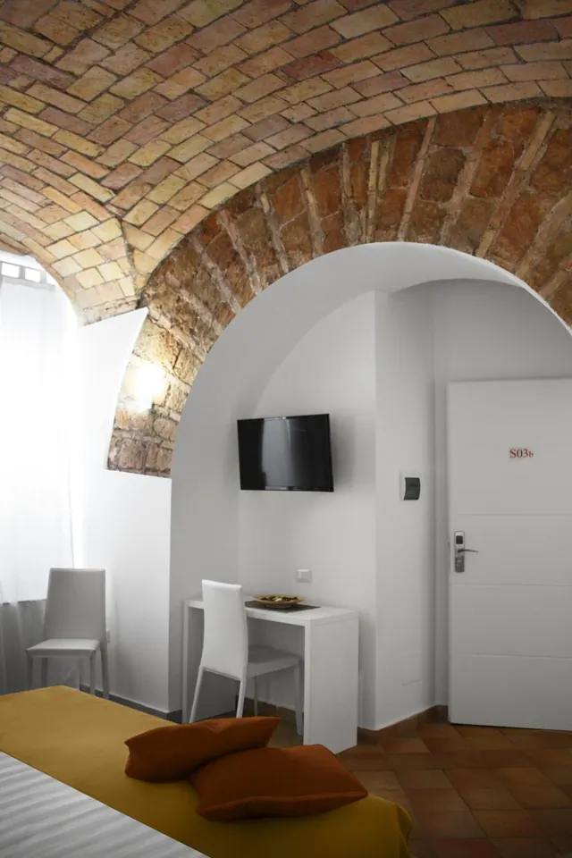 A bedroom with an exposed brick vaulted ceiling, a white arch, a wall-mounted television, a white desk and chair, and a bed with orange pillows in the foreground.