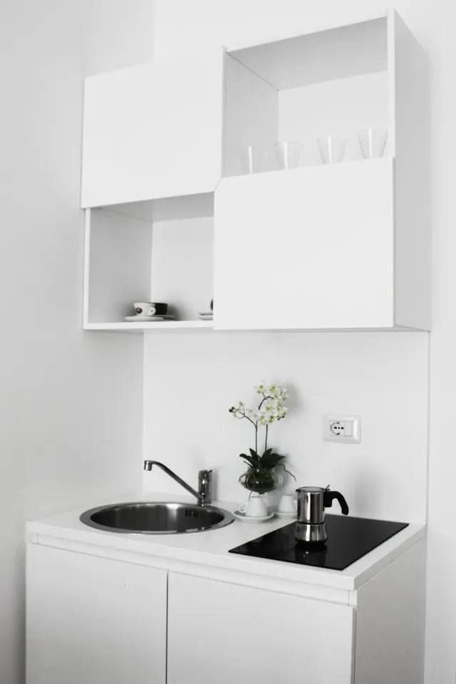 A small white kitchenette with a round steel sink, an induction hob, a moka pot, an orchid in a glass vase, and open and closed wall cabinets holding glasses and cups.