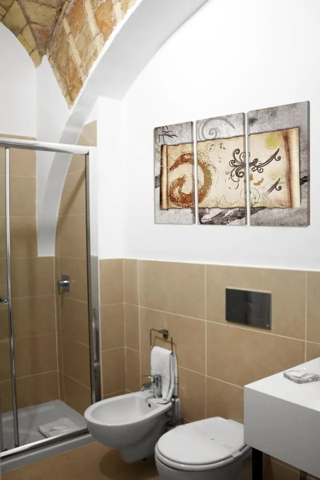 A bathroom with a glass-door shower, a wall-hung bidet and toilet, tan wall tiles, and a three-panel canvas on the wall.