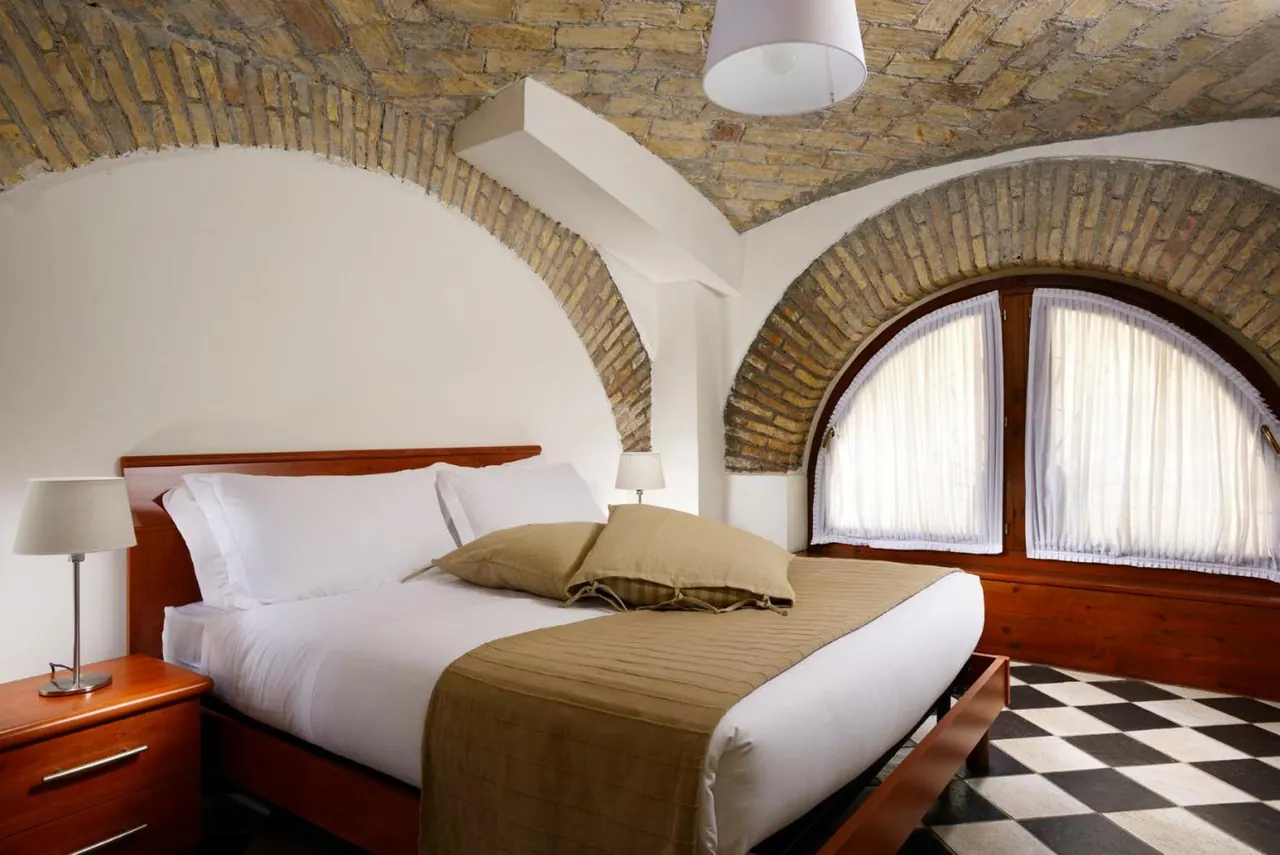 A double bed with white linen and a brown throw sits under a brick vaulted ceiling beside an arched window with sheer curtains.