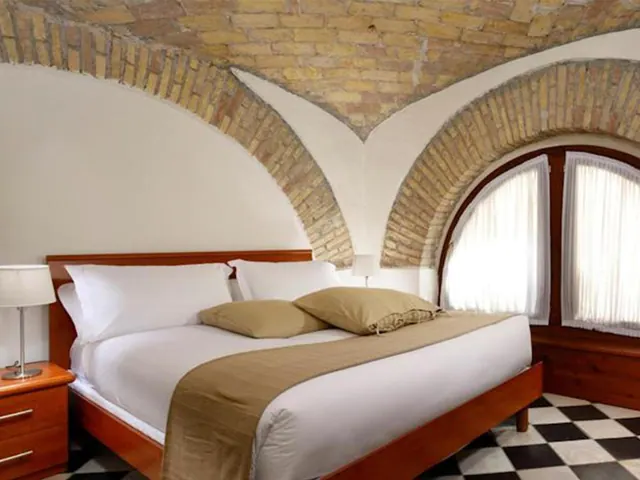 A double bed with white linen and a tan runner sits beneath a brick vaulted ceiling, beside a wooden nightstand and an arched window with sheer curtains.