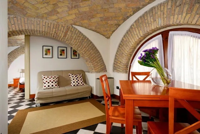 A living area with a beige sofa with polka-dot cushions, a wooden dining table with a vase of purple flowers, and a brick vaulted ceiling.