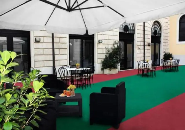 An outdoor terrace with a white umbrella, black chairs and tables, and a green and red painted floor beside a stone building with arched windows.
