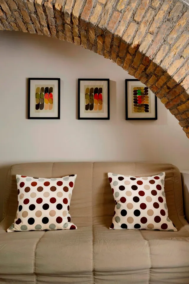 A beige sofa with two polka-dot cushions sits beneath an exposed brick arch, with three framed abstract prints on the wall above it.