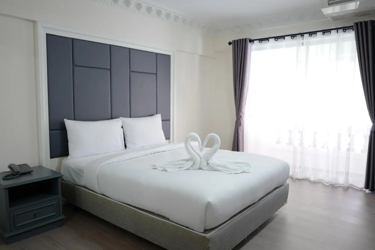A made king bed with a grey upholstered headboard and towel swans on the bedding, with a bedside table holding a telephone and sheer curtains over a large window.