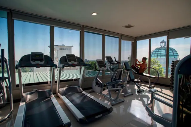 A gym with treadmills and exercise bikes in front of floor-to-ceiling windows overlooking the city, with one person using a bench on the right.