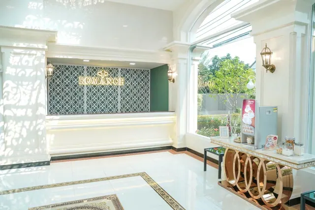 A lobby with a reception desk under a ROMANCE sign, a drink dispenser on a console table, and an arched window looking onto a garden with trees.