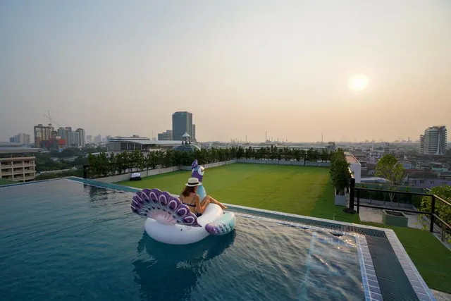 A woman in a sun hat sits on a peacock-shaped pool float in a rooftop infinity pool, with a lawn and city skyline beyond at sunset.
