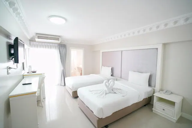 A bedroom with two white beds, a towel swan on the nearer bed, a wall-mounted television, a desk, and an air conditioner unit, with an open bathroom door visible beyond.