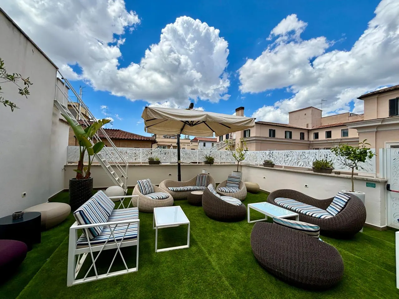 A rooftop terrace with artificial grass, wicker lounge seating with striped cushions, white metal chairs, and a cream parasol under a blue sky with clouds.