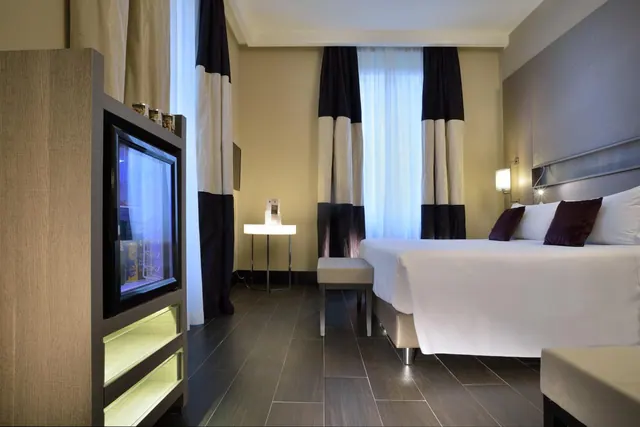 A hotel bedroom with a made bed with white linens and purple cushions, a television in a wooden cabinet with lit shelves, and a window with sheer curtains.