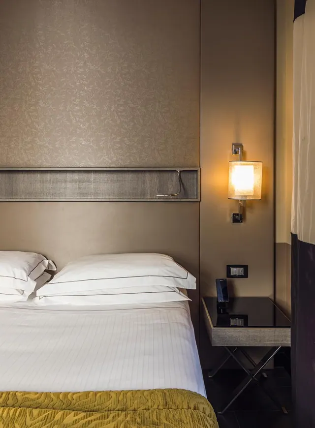 A double bed with white pillows sits beside a lit wall lamp and a black bedside table in a hotel room.