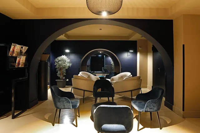 A dimly lit lounge with velvet armchairs, cream sofas, a potted plant and a large curved archway, with a magazine rack on the left.
