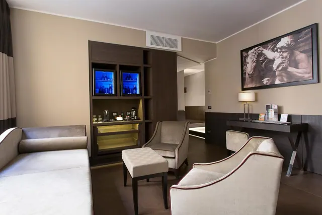 A hotel room seating area with a sofa, armchairs, an ottoman, a cabinet with blue-lit minibar fridges and a coffee machine, and a framed sculpture photograph on the wall.