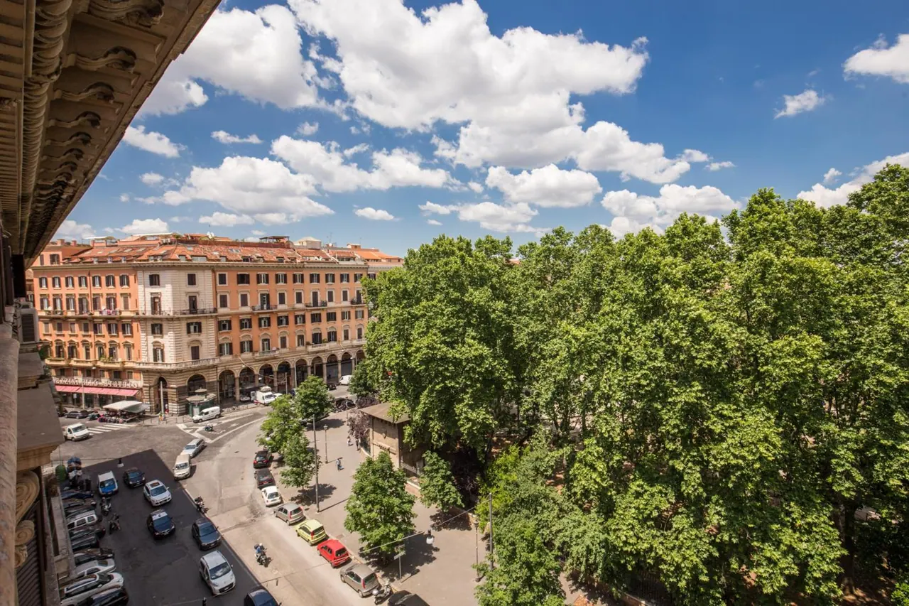 View from an upper floor over a tree-lined piazza with parked cars, a road and ochre apartment buildings under a partly cloudy blue sky.
