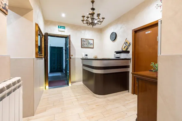 A hotel reception area with a curved dark reception desk, a printer on the counter, an ornate chandelier, a wall clock and an open door leading to a corridor.