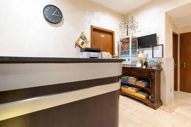 A hotel reception desk with a wall clock, a printer, a drinks fridge, a wooden sideboard with supplies and a wall-mounted television.
