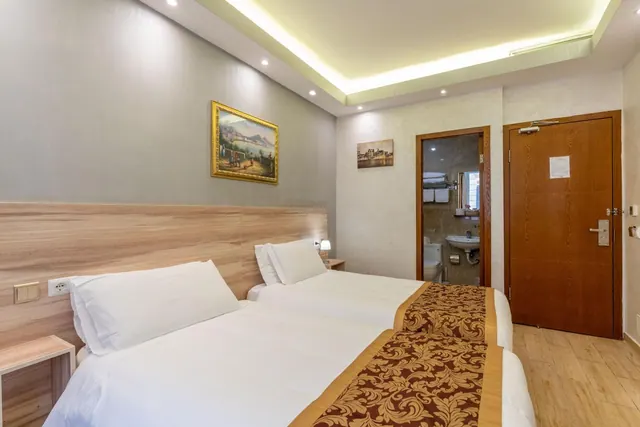 A bedroom with two single beds with white bedding, a framed painting on the wall, and an open door to a bathroom with a sink and toilet.