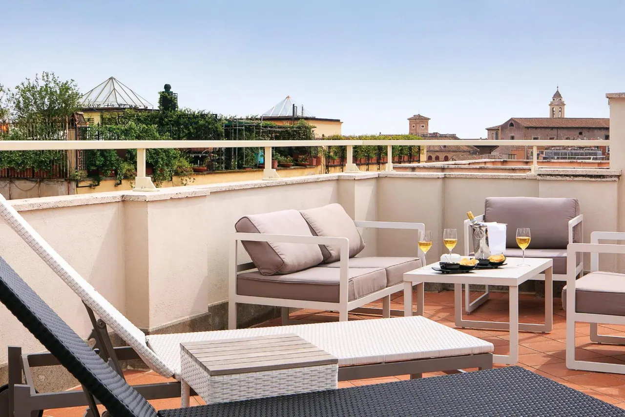A rooftop terrace with white lounge chairs, a sofa set and a coffee table holding two glasses of white wine, with rooftops and a church bell tower visible beyond the railing.