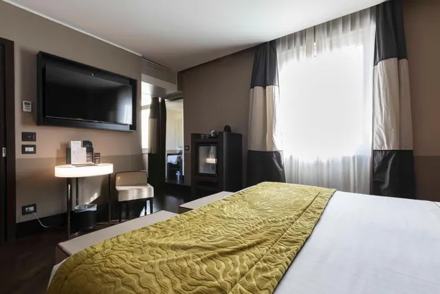 A hotel bedroom with a made double bed, a wall-mounted television, a round illuminated side table with a chair, and a dark fireplace unit beside a bright window with sheer and striped curtains.