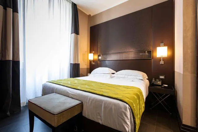 A double bed with white pillows and a yellow-green throw sits against a dark padded headboard, with an upholstered bench at the foot and wall lamps on each side.