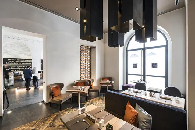 A dining area with set wooden tables, upholstered armchairs, dark pendant lights and a tall arched window, with a guest with luggage visible through a doorway to the lobby.