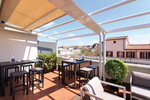 A rooftop terrace with a white pergola, high bar tables and stools, potted plants and lounge chairs, with rooftops and buildings visible beyond the railing.