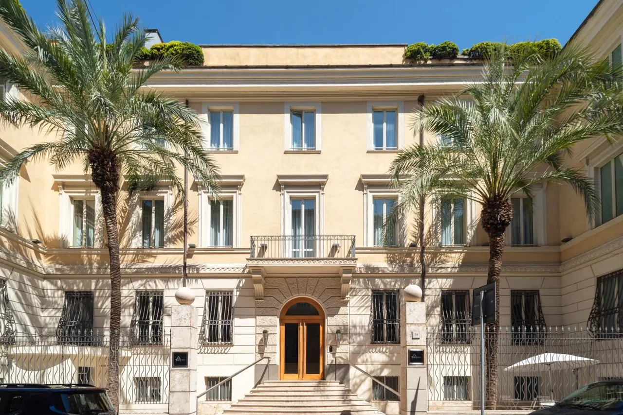 Front facade of a cream-coloured multi-storey building with two palm trees, an arched wooden entrance door at the top of a stone staircase, and iron railings along the street.