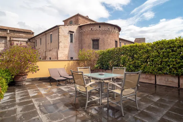A rooftop terrace with a glass-topped table, four chairs, two sun loungers, potted plants and a tall hedge, with an old brick building behind.