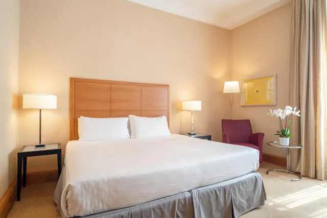 A bedroom with a made double bed, two bedside lamps, a red armchair, and a potted white orchid beside floor-length curtains.