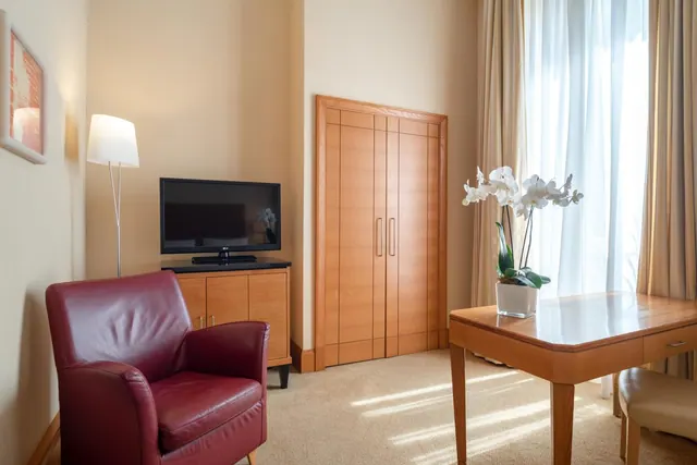A living area with a red leather armchair, a flat-screen television on a wooden cabinet, a floor lamp, a wooden wardrobe and an orchid on a wooden desk by a window with sheer curtains.