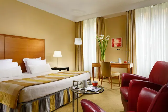A hotel bedroom with a double bed, a desk by the tall curtained windows, and red leather armchairs around a glass coffee table.