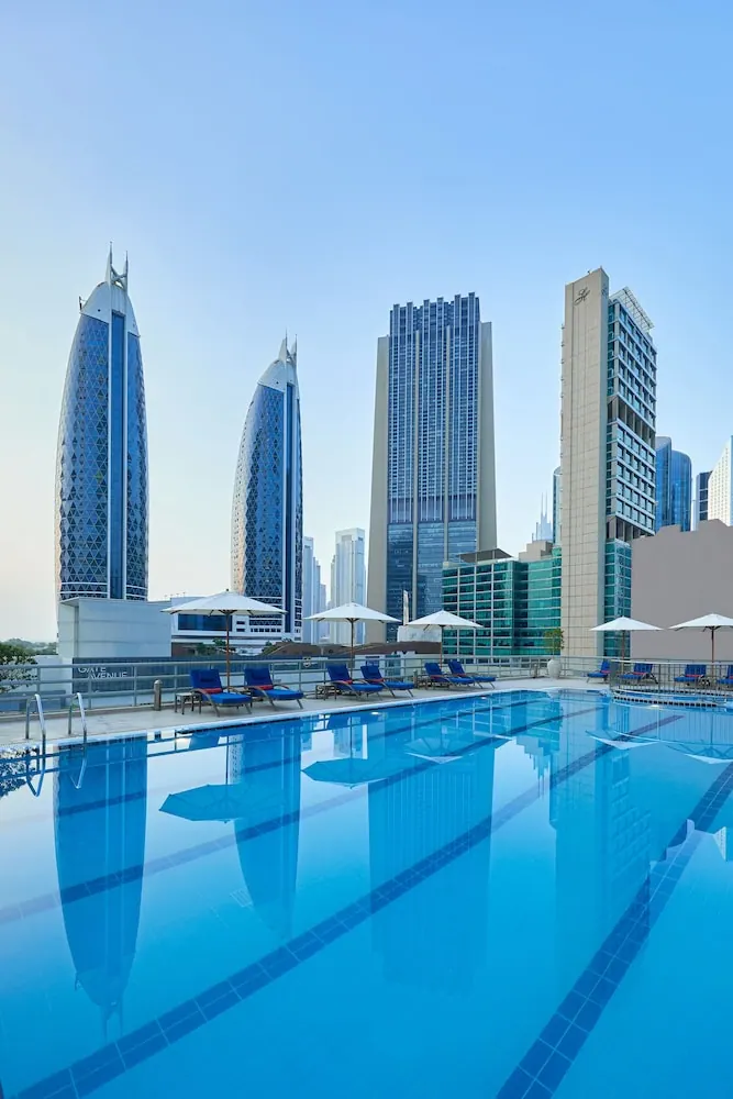 A rooftop swimming pool with blue loungers and white parasols, with high-rise towers visible beyond the railing.