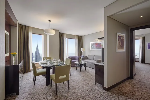 A suite living and dining area with a round dining table set for two, a grey sofa, and tall windows showing a tall spired tower and sky outside.