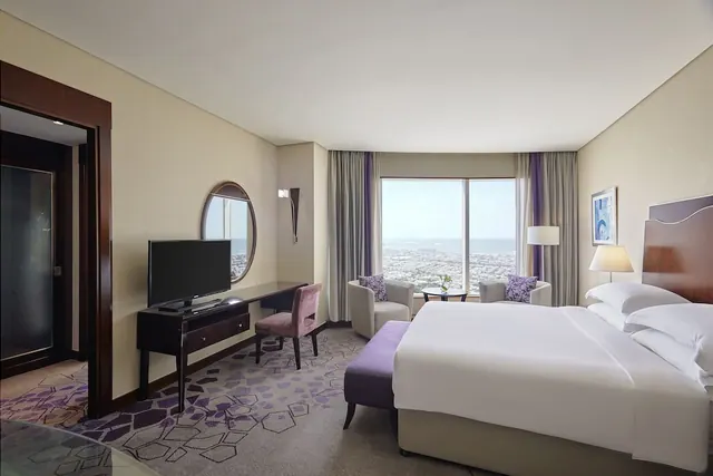 A hotel bedroom with a king bed, a desk with a television, two armchairs, and a large window overlooking the sea and low buildings.