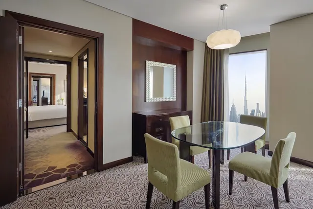 A dining area in a hotel suite with a glass oval table and green upholstered chairs, with the Burj Khalifa visible through the window and an open doorway to a bedroom beyond.