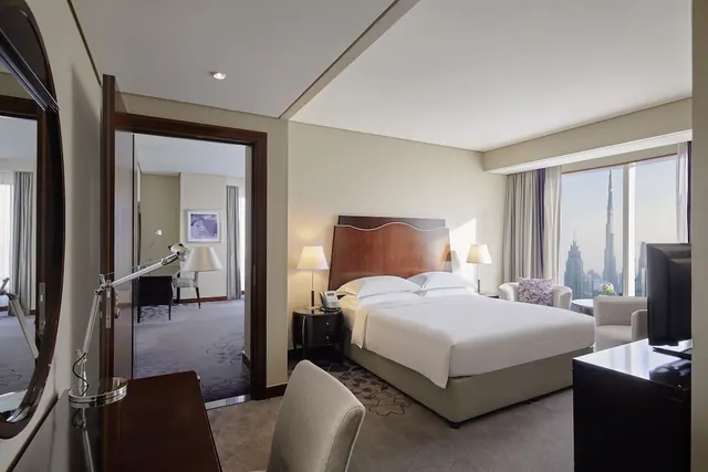 A bedroom with a made king bed, an armchair seating area, a wall-mounted television, and a window showing the Burj Khalifa and the city skyline.