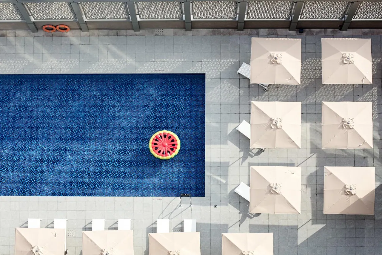 Overhead view of a rectangular blue-tiled pool with a watermelon-shaped float, cream square umbrellas and white sun loungers on grey pavers.