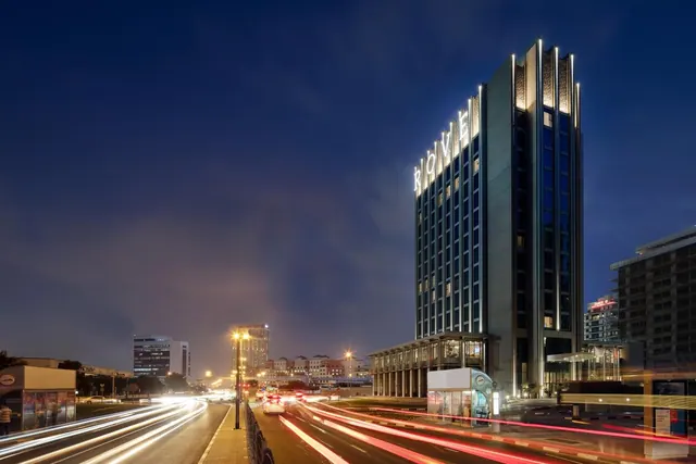 A tall illuminated hotel tower with the ROVE sign on its upper section stands beside a road with light trails from passing traffic at night.